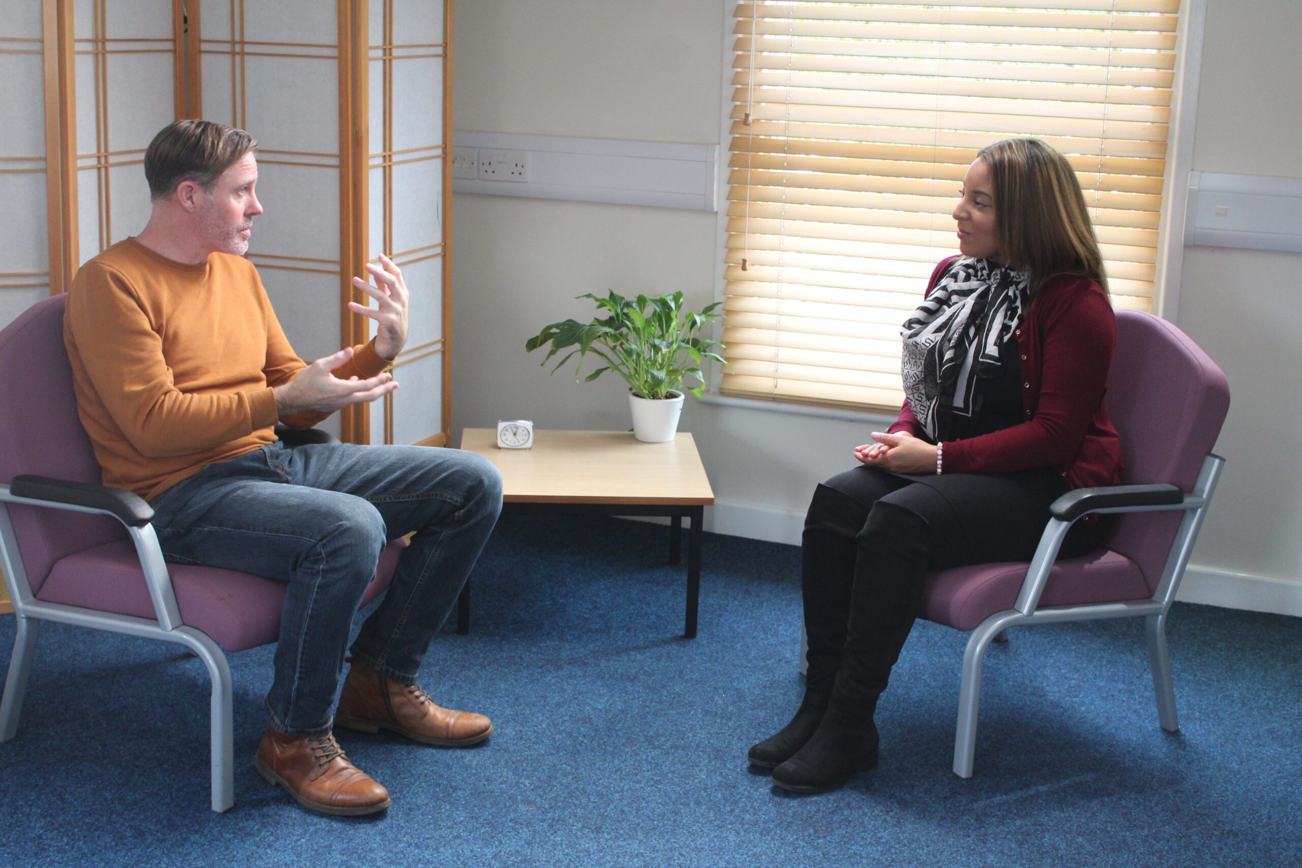 A man and a woman sit in a room in a counselling-style setting. The man is talking expressively to the woman.