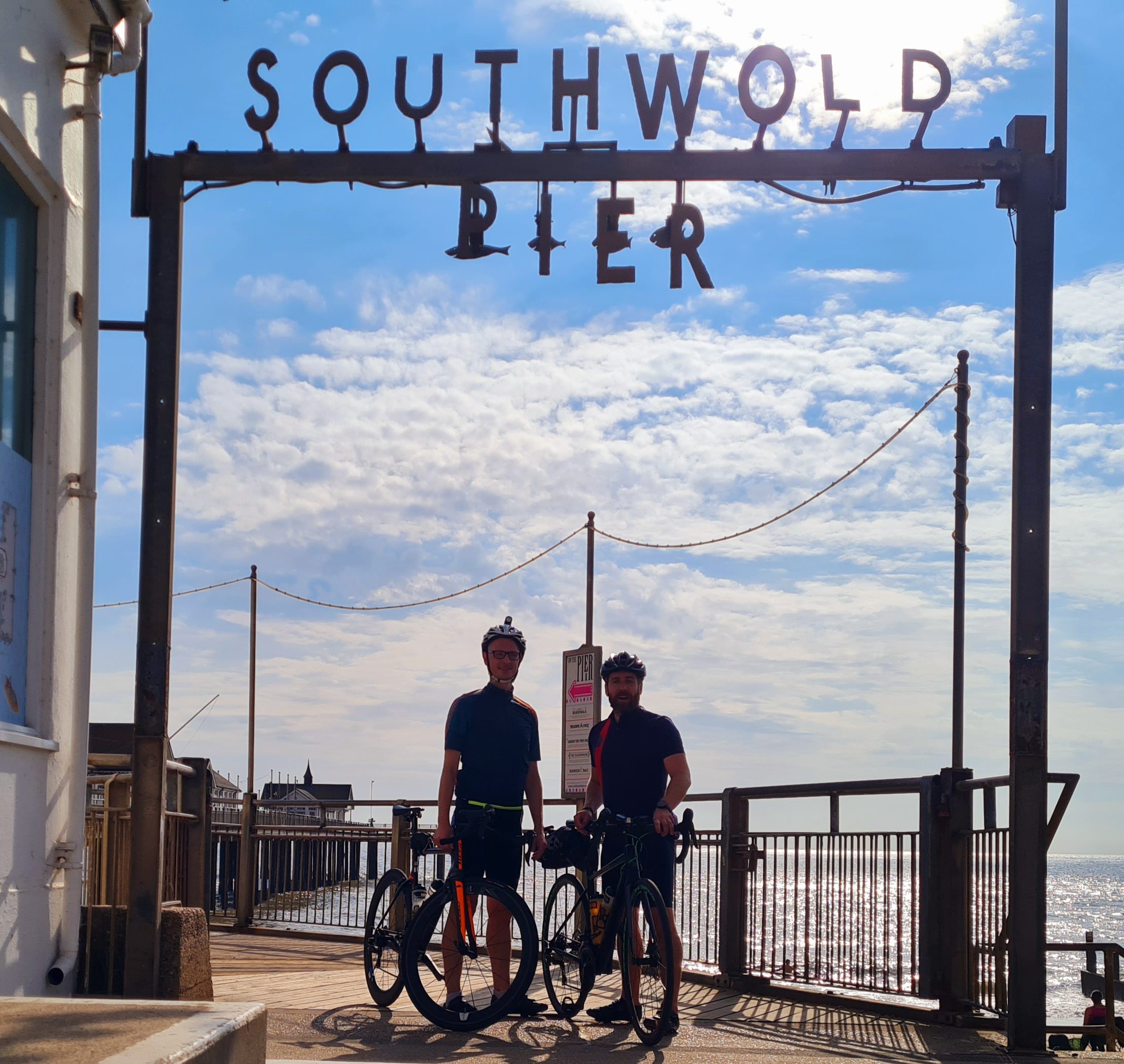Two people on bicycles in front of Southwold Pier with sunshine and clouds behind them