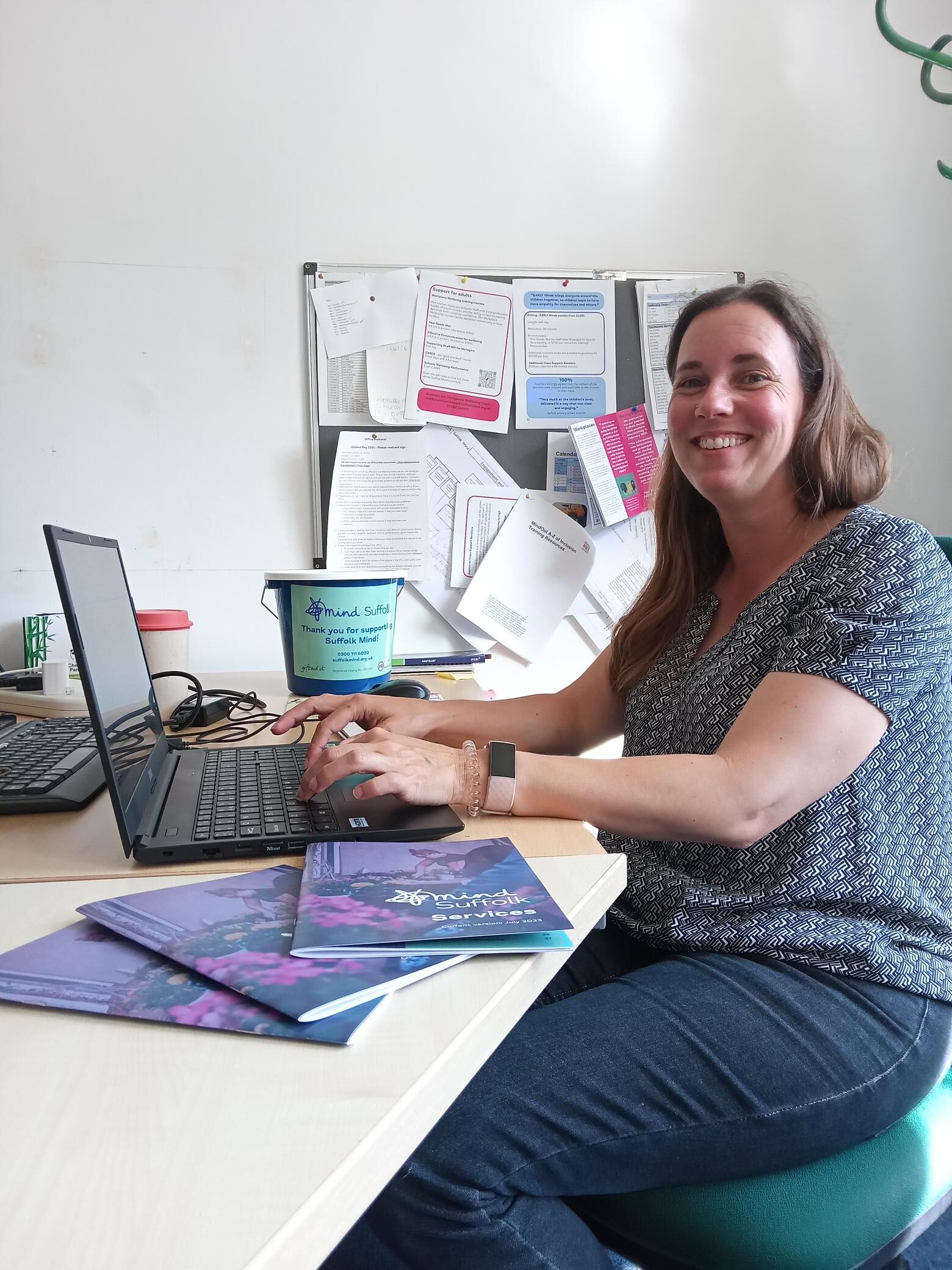 A woman sits at a desk in an office environment, with her hands on the laptop, smiling to the camera.