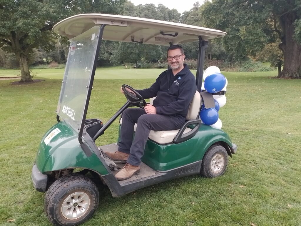 A man smiles at the camera while sitting in a golf caddy. 'Suffolk Mind' is written on his fleece. 