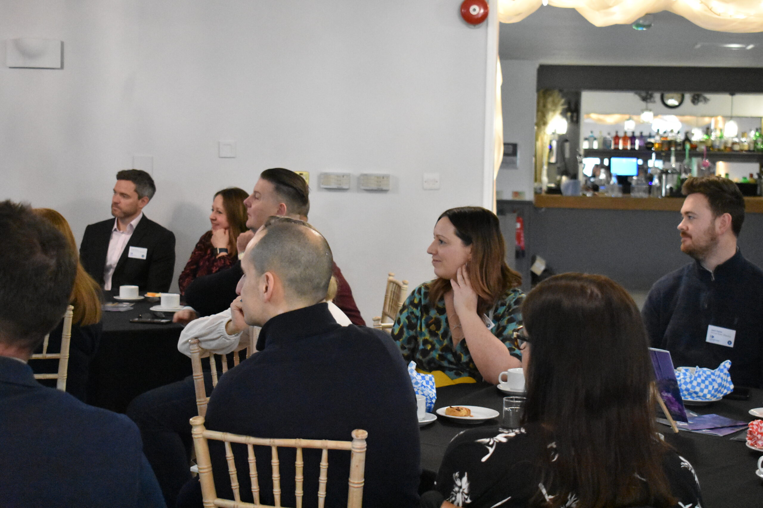 A group of people sit at a table in a conference setting, all paying attention to a speaker who is out of shot.