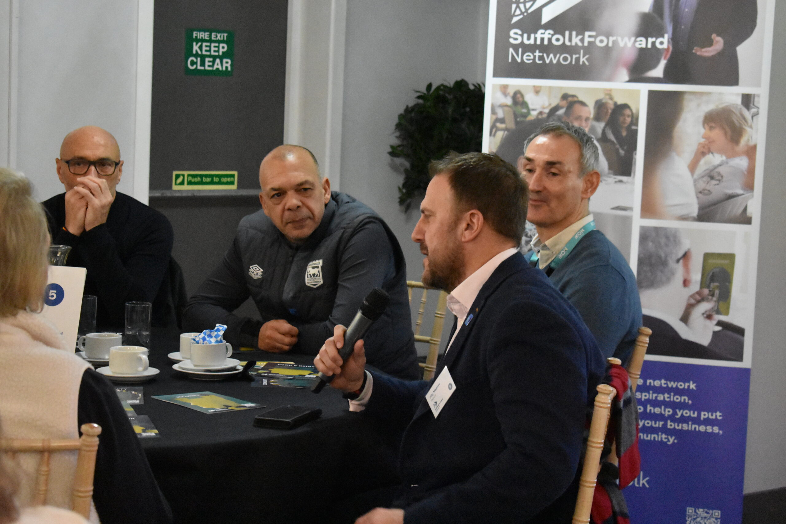 In a business networking setting, a man speaks into a microphone while two other men at the table pay attention.