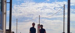 Two people on bicycles in front of Southwold Pier with sunshine and clouds behind them