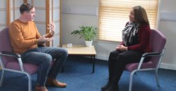 A man and a woman sit in a room in a counselling-style setting. The man is talking expressively to the woman.