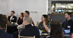 A group of people sit at a table in a conference setting, all paying attention to a speaker who is out of shot.