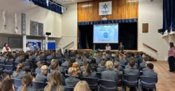 Picture shows an assembly of secondary schoolchildren in a school hall. The children are seated in rows looking towards the stage where a presentation is being delivered.
