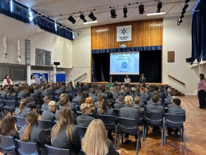 Picture shows an assembly of secondary schoolchildren in a school hall. The children are seated in rows looking towards the stage where a presentation is being delivered.