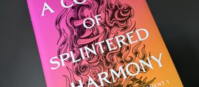 Hand holding the hardback copy of A Court of Splintered Harmony against a dark grey wall