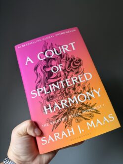 Hand holding the hardback copy of A Court of Splintered Harmony against a dark grey wall