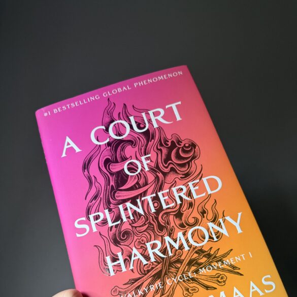 Hand holding the hardback copy of A Court of Splintered Harmony against a dark grey wall